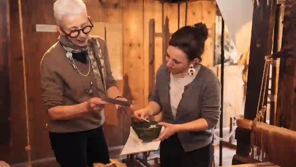 Inmitten des warmen Ambientes des Regionalmuseums Chüechlihus in Langnau teilen zwei Frauen ein Lächeln, während sie handwerkliche Fertigkeiten und Traditionen austauschen, umgeben von der Gemütlichkeit historischer Wände.