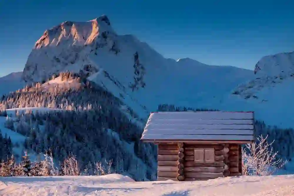 Das Bild zeigt eine schneebedeckte Blockhütte im Vordergrund, im Hintergrund die imposanten, sonnenbeschienenen Gipfel des Gantrisch-Gebirges. Die friedliche Szene strahlt Ruhe und die Stille einer frischen Winterlandschaft aus.