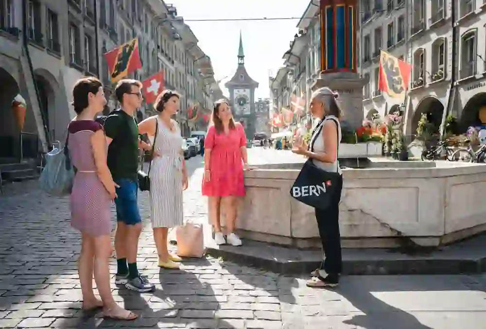 Auf dem Bild ist eine Gruppe von Menschen zu sehen, die an einer Stadtführung teilnehmen. Eine Führerin mit einer Umhängetasche, auf der "Bern" steht, erklärt etwas. Im Hintergrund sieht man die Kramgasse mit der Zytglogge (Zeitglocke). Es handelt sich um eine Stadtführung in der Stadt Bern.