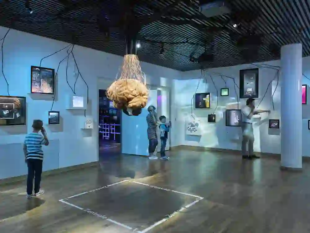 Eine dynamische Ausstellung im Museum mit einem interaktiven, lehrreichen Ansatz. Besucher jeglichen Alters sind vertieft in multimediale Displays, umgeben von farbenfroher Beleuchtung und kreativen Exponaten.