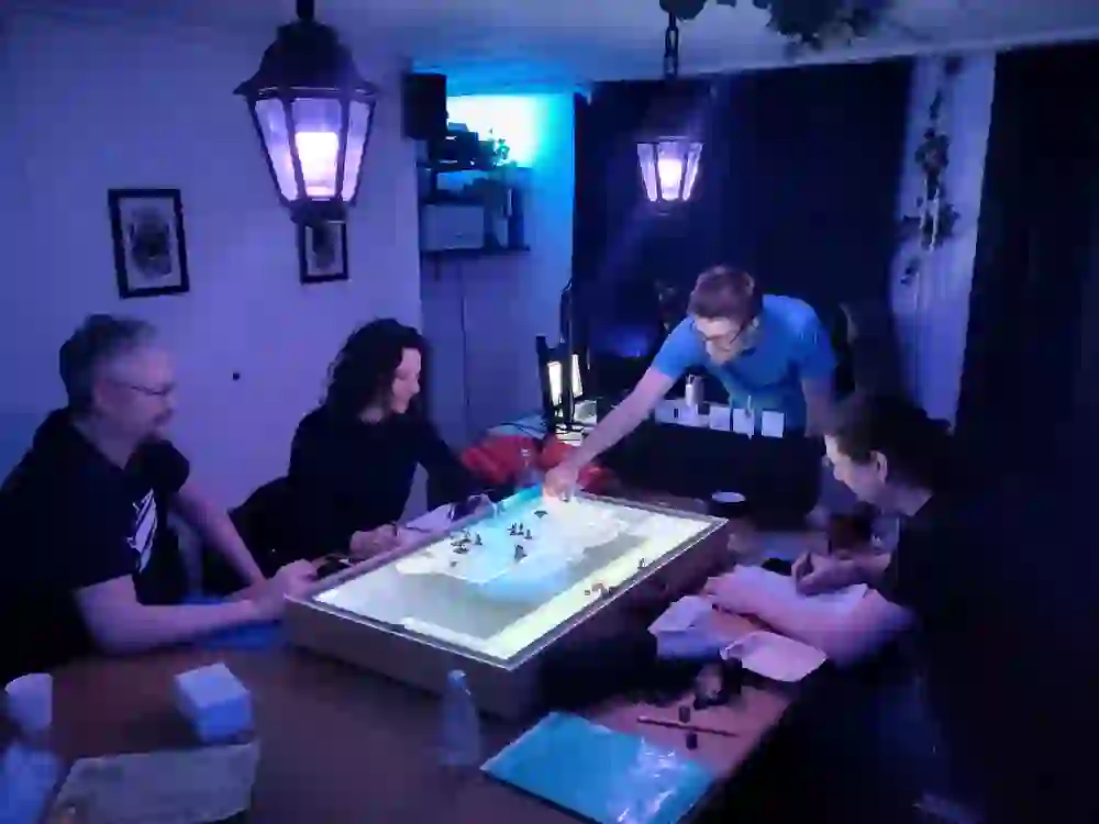 Mehrere Personen sitzen um einen beleuchteten Spieltisch und spielen gemeinsam ein Pen-&-Paper-Rollenspiel. Würfel, Figuren und Notizen liegen aus, die Atmosphäre ist konzentriert und zugleich locker.