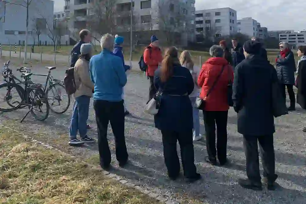Un gruppo interessato ascolta una visita guidata; gesti vivaci, incontro rilassato in un clima soleggiato, biciclette sullo sfondo.