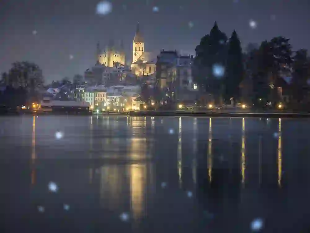 Das Bild zeigt eine nächtliche Winterlandschaft in Thun, Schweiz, mit schneebedeckten Gebäuden, darunter ein beleuchtetes Schloss. Die Lichter spiegeln sich im ruhigen Wasser, während Schneeflocken sanft fallen, was eine friedliche und magische Atmosphäre schafft.