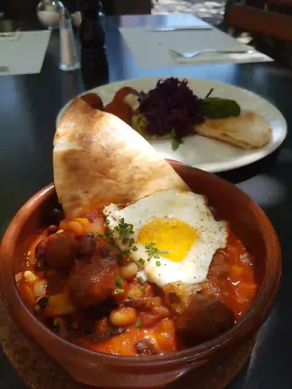 A rustic, hearty dish with a fried egg in a terracotta pot, served with bread and red cabbage, invites you to enjoy a cosy feast in a sunny ambience.