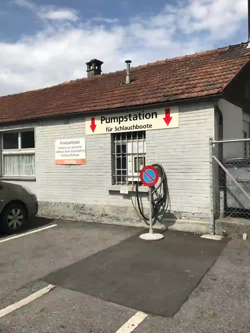 Auf dem Foto ist die Pumpstation in Schwäbis für Schlauchboote zu sehen, die sich an einer weissen Hauswand neben einem Parkplatz befindet. Rote Pfeile weisen auf die Pumpstation hin. Im Vordergrund steht eine Rolle mit einem Schlauch, Teil der Pumpausrüstung. Es herrscht ein pragmatischer, funktionaler Eindruck – ein Ort, der Aktivitäten auf dem Wasser vorbereitet.