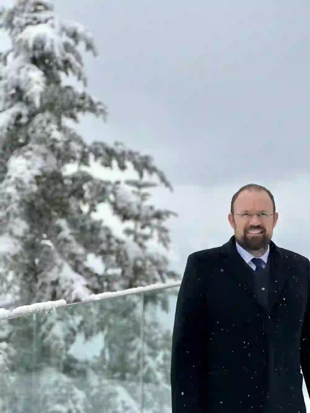 L'image montre un homme vêtu d'un élégant manteau sombre devant un paysage hivernal de sapins enneigés. Son sourire et son attitude détendue créent une atmosphère chaleureuse et conviviale malgré le froid ambiant.