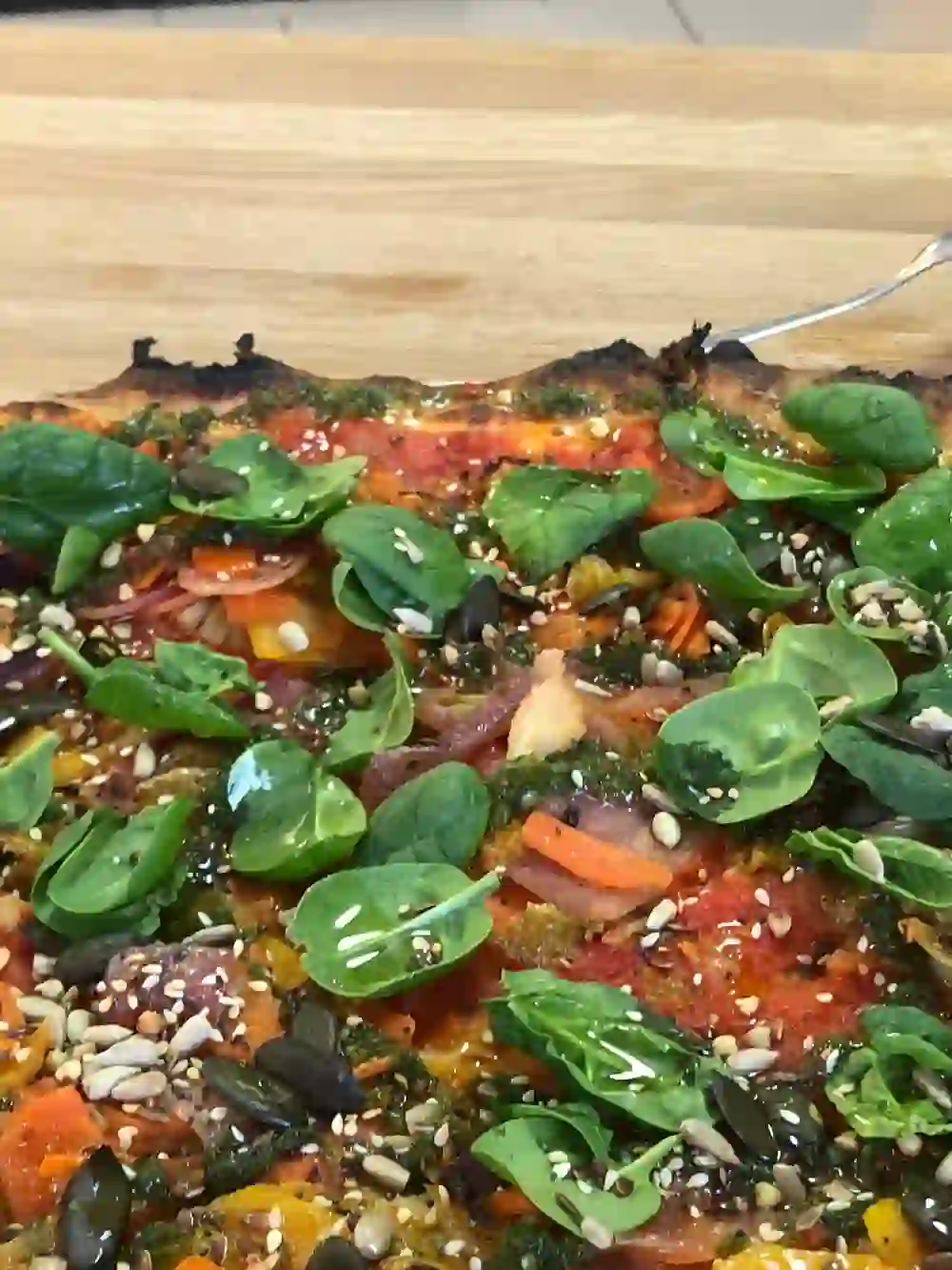 Das Bild zeigt eine appetitlich belegte Pizza, reich garniert mit frischem Spinat, bunten Paprikastreifen und einer Vielzahl von Samen. Die lebendigen Farben und frischen Zutaten vermitteln Frische und Geschmacksvielfalt.