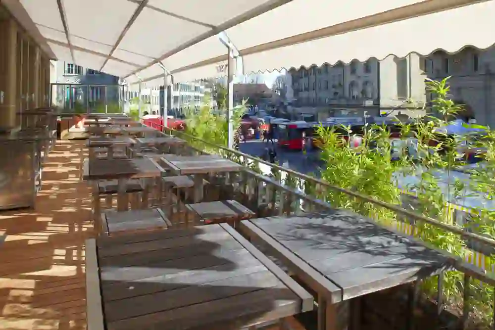The picture shows the outdoor terrace of the Mishio Vatterland Bern restaurant, covered by a large awning. There are several wooden tables and benches. The terrace is decorated with plants and offers a view of a busy street with passing buses and pedestrians. It looks like a pleasant place to eat outside.