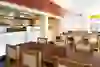 The picture shows the interior of the Mishino Vatterland Bern restaurant with modern furnishings. Several wooden tables and matching chairs with white seats can be seen. In the background is an open kitchen with a person who appears to be working in the kitchen. The colour scheme is neutral with white walls and an orange accent on the ceiling. Large windows let in natural light.