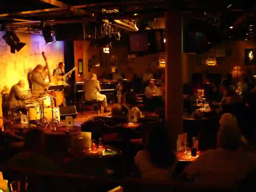 The scene shows a lively concert at Marians Jazzroom in Bern. Between the subdued light and the shadows, the musicians on stage cast vivid silhouettes. Audience members sit at tables, enveloped in the soft lighting as they listen to the sounds of jazz and the rhythm of the drums. It is a place of gathering and enjoyment, where music unites people and creates a nostalgic, almost timeless atmosphere.