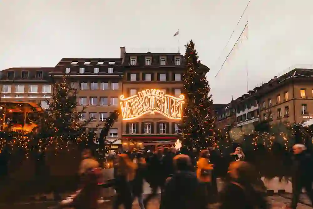 Das Bild zeigt den festlich beleuchteten Berner Weihnachtsmarkt auf dem Waisenhausplatz. Menschen strömen vorbei, in warme Jacken gehüllt, während die Lichter eine magische und einladende Atmosphäre schaffen. Die Stimmung ist lebendig und voller Vorfreude.