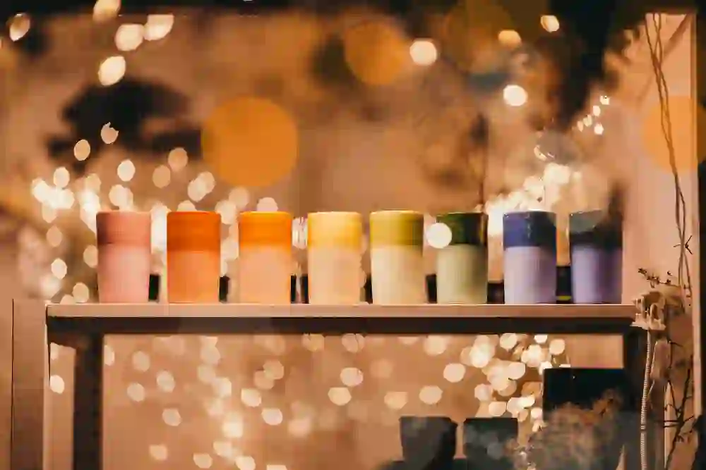 The picture shows a row of colourful, handmade mugs on a shelf, illuminated by warm fairy lights in the background. The soft lighting and bright colours create a cosy, festive atmosphere that invites you to linger.