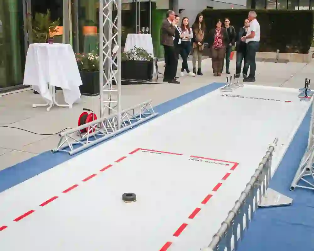 Das Bild zeigt eine Eisstockschießbahn auf einer Terrasse. Im Vordergrund ist die schwarze Daube zu sehen, und mehrere Eisstöcke stehen am Ende der Bahn. Im Hintergrund steht eine Gruppe von Personen, die dem Geschehen zuschauen.