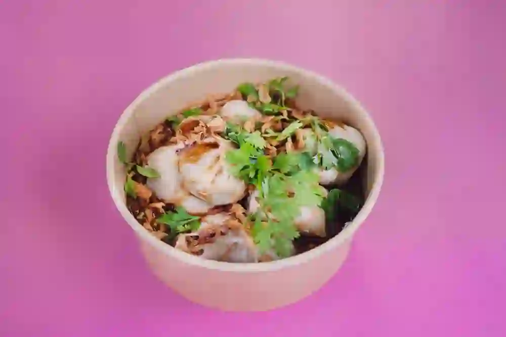 Das Bild zeigt eine Schale mit saftigen Momos, garniert mit frischem Koriander und knusprigen Zwiebeln. Der lebendige, pinke Hintergrund betont die Farbenfreude des Gerichts, was eine fröhliche und genussvolle Stimmung vermittelt.