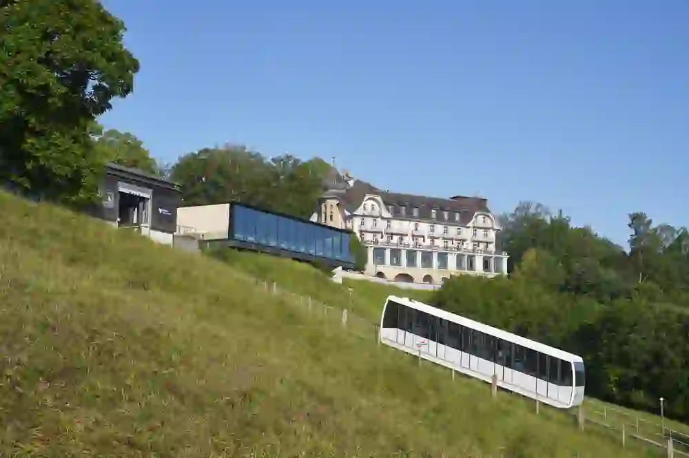 L'image montre une scène idyllique avec un funiculaire moderne qui se déplace sur une pente verdoyante à côté d'un bâtiment classique et élégant. Le mariage harmonieux de la tradition et de la modernité dégage une impression de calme et de progrès.