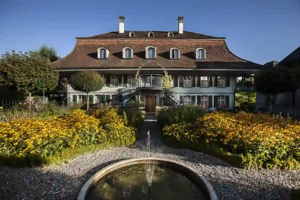 La foto mostra l'idilliaco giardino del Romantikhotel Bären Dürrenroth. Aiuole fiorite e una fontana gorgogliante di fronte all'edificio storico creano un'atmosfera tranquilla e pittoresca.