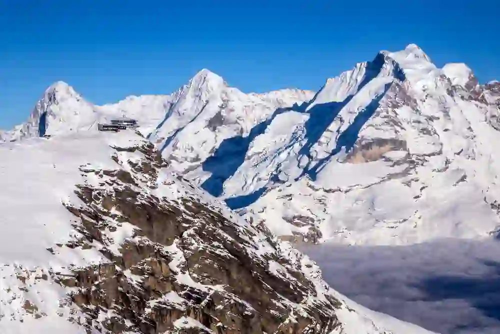 L'immagine mostra le cime innevate delle Alpi svizzere con la stazione montana dello Schilhorn su una sporgenza, sotto un cielo azzurro e limpido e sopra una coltre di nuvole.