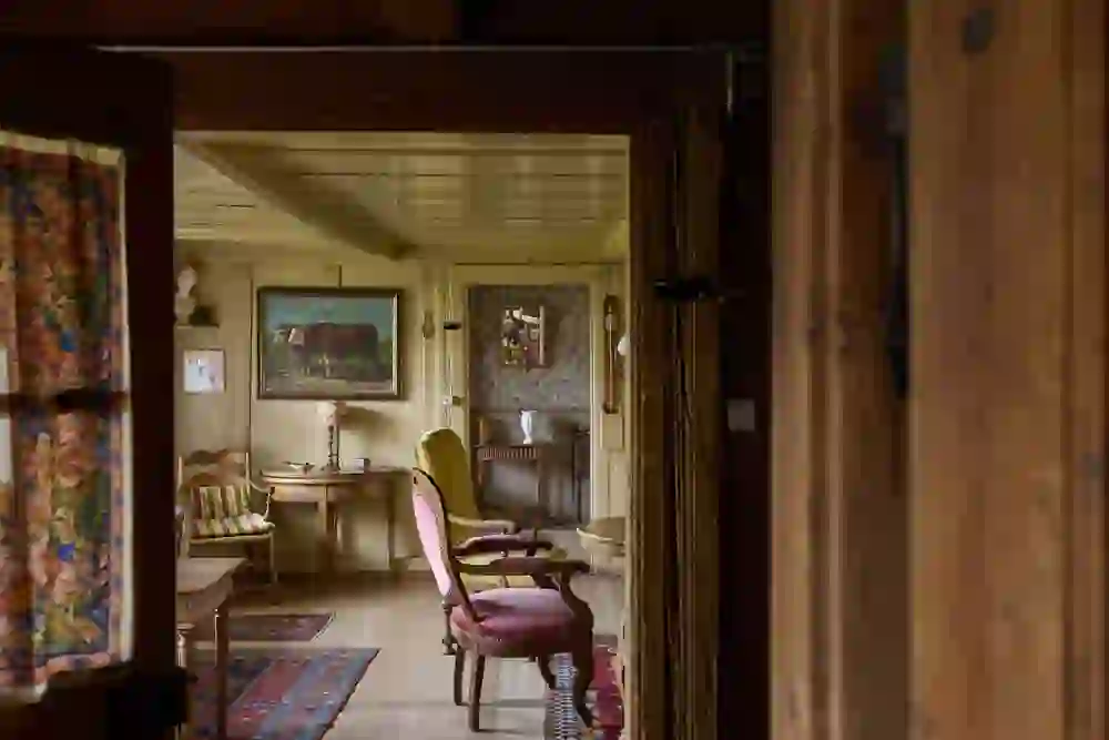 L'immagine mostra un soggiorno arredato con amore in una casa tradizionale con elementi in legno. Mobili antichi, tappeti colorati e dettagli decorativi come un dipinto di mucche e una carta da parati ornata conferiscono all'ambiente un'atmosfera calda e nostalgica.