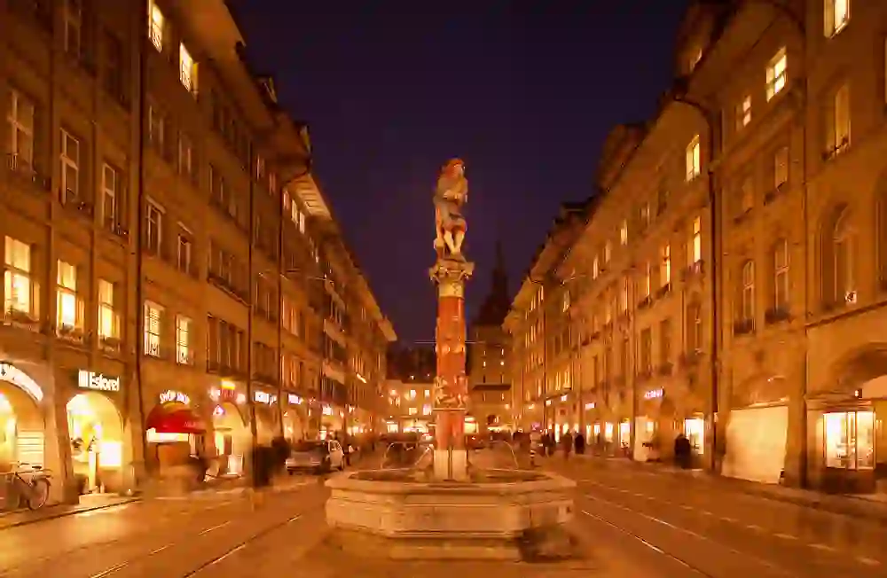 Auf dem Bild ist der Pfeiferbrunnen in Bern bei Nacht zu sehen, eine belebte Straße mit Geschäften im Hintergrund und die warme Beleuchtung verleiht der Szene eine einladende Atmosphäre.