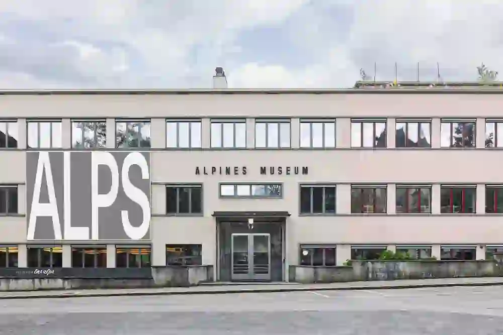 Das Bild zeigt die Fassade des Alpinen Museums in Bern. Das Gebäude ist modern und schlicht gestaltet, mit großen Fenstern und einer klaren, geometrischen Architektur. Über dem Eingang steht der Schriftzug „ALPINES MUSEUM“, während auf der linken Seite ein großes Banner mit der Aufschrift „ALPS“ angebracht ist. Im unteren Bereich ist das Restaurant „les alps“ sichtbar. Die Fassade kombiniert neutrale Farbtöne mit einem leichten Rosaton. Auf dem Dach sind kleine Fahnen zu erkennen, die dem Gebäude eine lebendige Note verleihen. Die Szene vermittelt einen professionellen und einladenden Eindruck.