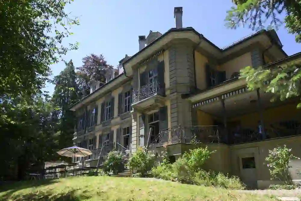 Die Villa Stucki erstrahlt an einem sonnigen Tag inmitten eines grünen Gartens. Das historische Gebäude mit seinen eleganten Balkonen, blühenden Sträuchern und schattigen Sitzplätzen verströmt eine ruhige, einladende Atmosphäre – ein Ort für Genuss und Entschleunigung.