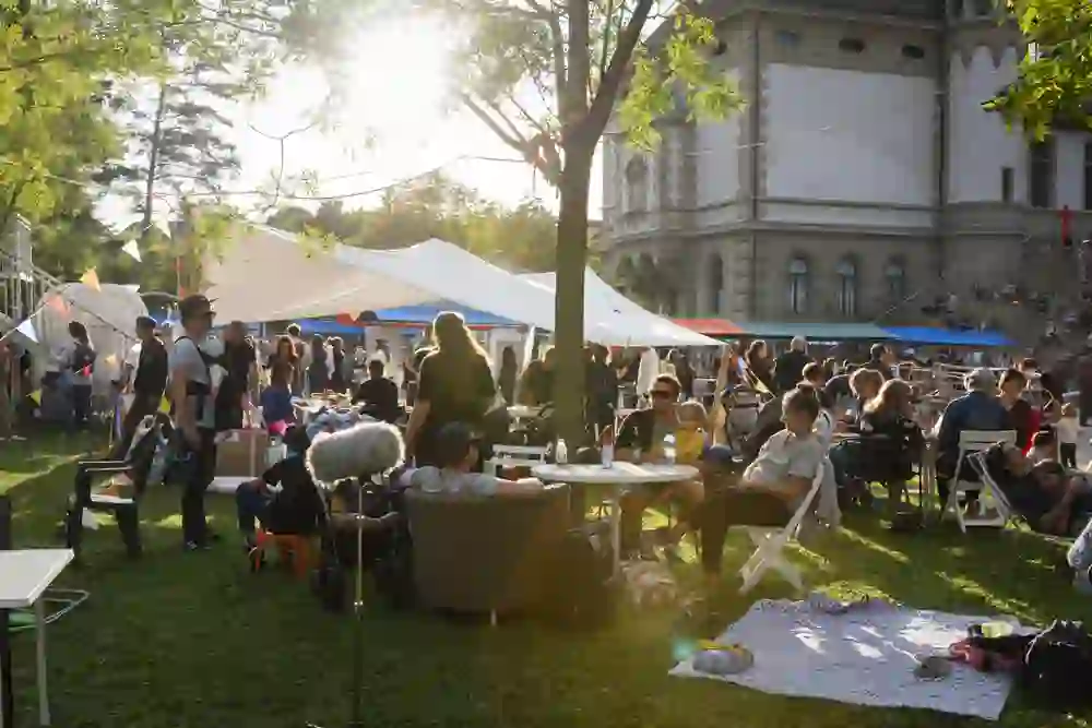Auf dem Bild ist eine belebte Outdoor-Veranstaltung zu sehen. Im Vordergrund sitzen und stehen Menschen an Tischen, die sich unterhalten und entspannen. Einige Menschen haben Kinder dabei. Im Hintergrund gibt es Zelte und bunte Wimpelketten, die eine fröhliche Atmosphäre schaffen. Die Veranstaltung findet auf einer Rasenfläche vor einem großen, historischen Gebäude statt. Die Sonne scheint, und es herrscht eine freundliche, sommerliche Stimmung.