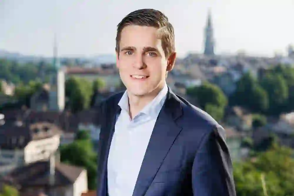 A self-confident man in a suit smiles in front of a cityscape. His gaze is friendly, the atmosphere professional.