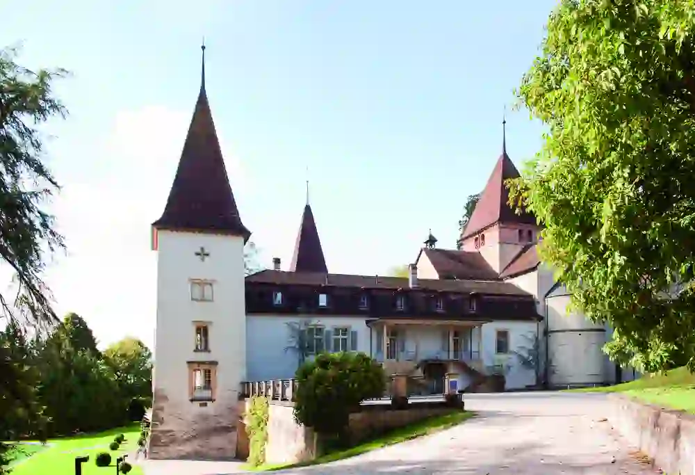 Das Bild zeigt Schloss Münchenwiler, umgeben von üppigem Grün. Die majestätische Architektur und die sonnige Atmosphäre laden zu einer ruhigen Entdeckungsreise in historischer Umgebung ein.