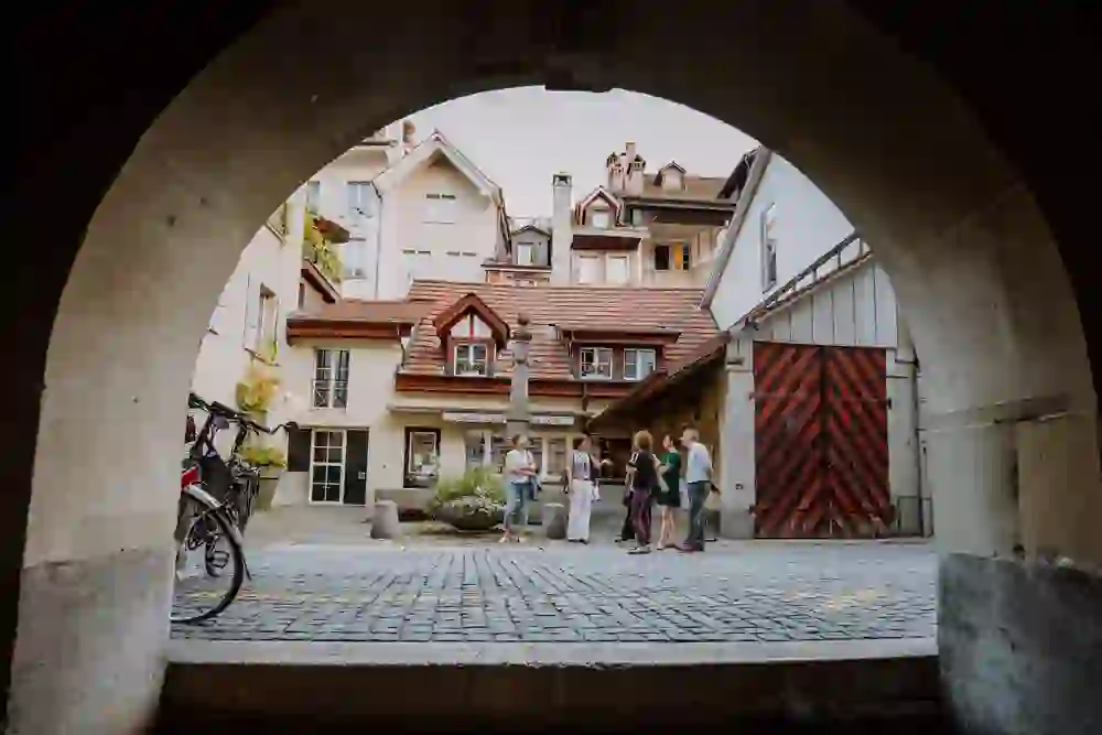 Durch ein Torbogen erblickt man eine Gruppe, die inmitten eines charmanten Berner Hofs steht. Sie sind in ein Gespräch vertieft, und die entspannte Körpersprache zeigt ihr Interesse an der Erzählung der Führerin. Das historische Ambiente und das sanfte Licht verleihen der Szene eine idyllische Ruhe.