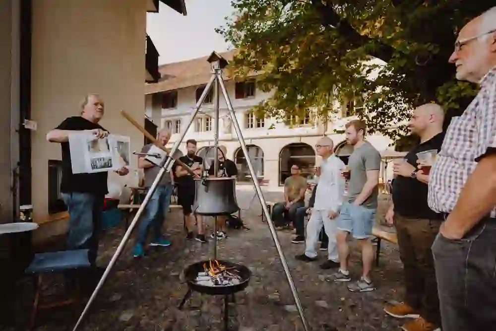 Das Bild zeigt eine gesellige Runde von Personen unterschiedlichen Alters um einen grossen Kessel, der über einem offenen Feuer hängt. Ein Mann präsentiert ein Bild, während die anderen entspannt zuhören und Getränke halten, was auf eine gemütliche Atmosphäre bei einem Freiluft-Event hinweist.