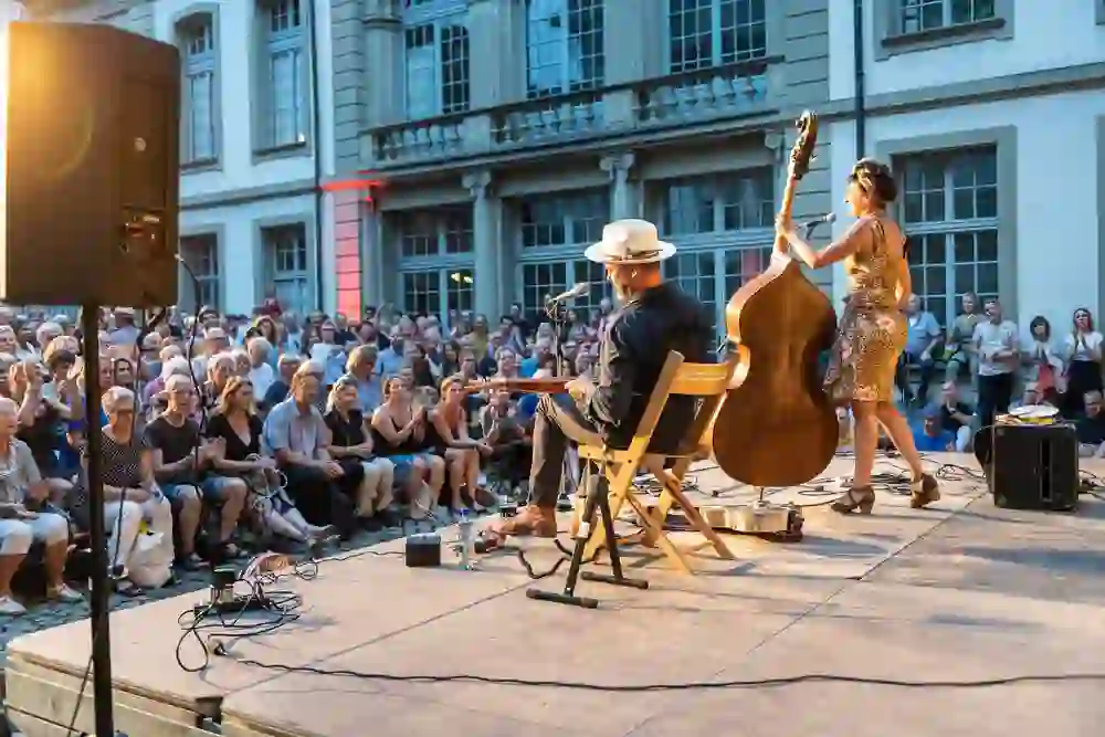 Ein Musikerduo begeistert das Publikum beim Buskers Festival in Bern. Der Gitarrist sitzt, die Kontrabassistin steht dynamisch, beide vor einer erfreuten Menschenmenge, die die sommerliche Abendluft und die lebendige Musik unter freiem Himmel geniesst. Die Stimmung ist ausgelassen und herzlich.