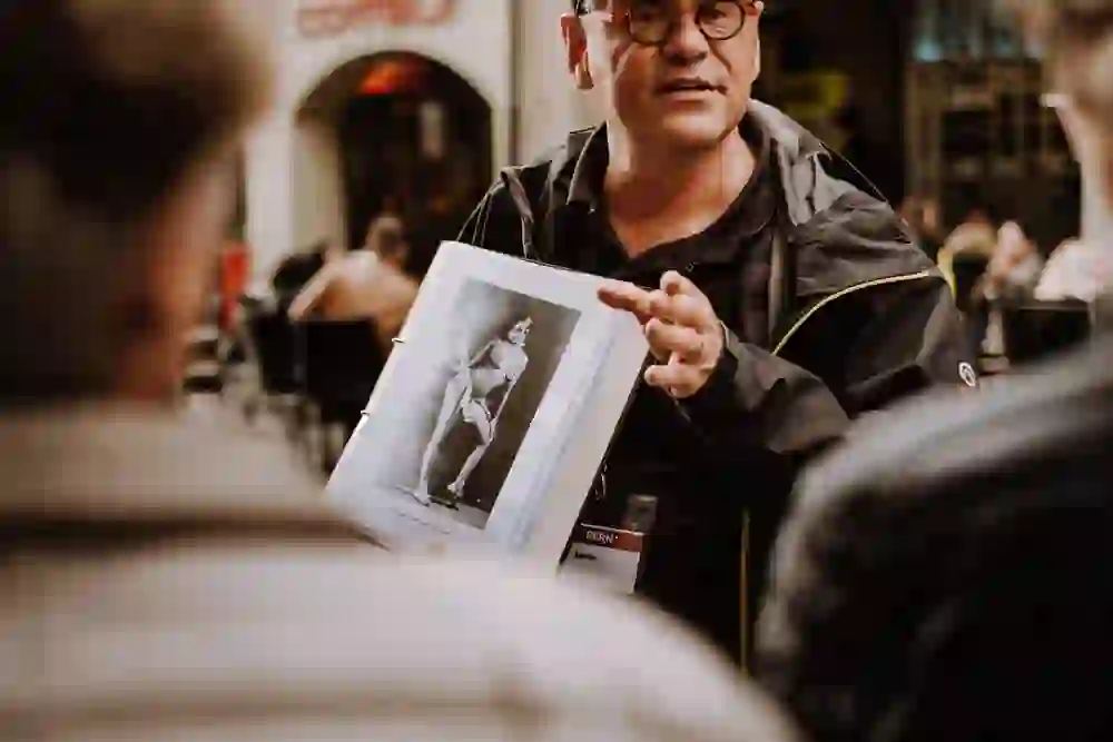 Auf diesem Bild sieht man einen Mann, der ein Fotoalbum oder eine Mappe hält und darauf zeigt, während er sich an eine Gruppe wendet. Seine Gestik wirkt erklärend und engagiert, vielleicht erläutert er historische Details oder Anekdoten über die Stadt Bern. Die Szene spielt vor einem Café, was auf eine lebhafte und gemütliche Atmosphäre schliessen lässt.