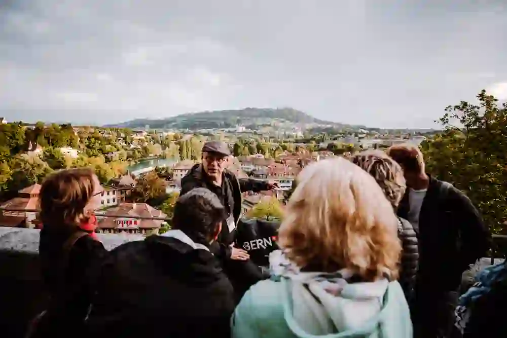 Ein Reiseführer, erkennbar an seinem Badge, erzählt einer Gruppe von Zuhörern, die ihm interessiert und aufmerksam lauschen, etwas über die Stadt Bern, die im Hintergrund zu sehen ist. Die Szene wirkt lebhaft und ist gefüllt mit einer Atmosphäre des Lernens und der Neugier.