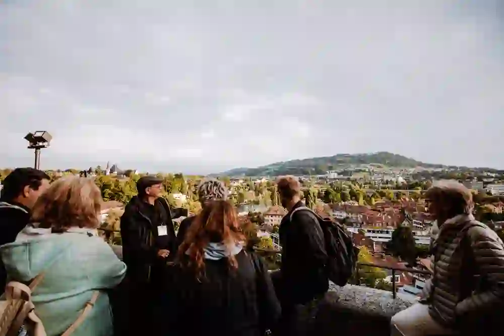 Das Foto zeigt eine Gruppe von Touristen, die von einem Aussichtspunkt aus auf die Dächer der Stadt Bern. Das Panorama zeigt eine malerische Ansicht mit Bäumen und traditioneller Architektur im Hintergrund. Die Gruppe steht entspannt beieinander und scheint die Aussicht zu genießen, mit einem Reiseleiter, der auf interessante Punkte hinweist. Die Atmosphäre wirkt ruhig und beschaulich.