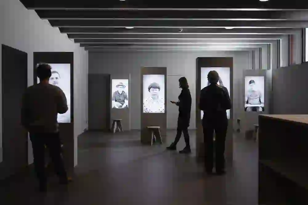 Auf dem Bild ist ein Ausstellungsraum des Schweizerischen Blindenmuseum mit großformatigen Porträtfotografien zu sehen. Personen betrachten die Werke, die auf einzelnen Stelen beleuchtet präsentiert werden. Die Stimmung wirkt ruhig und reflektierend, während die Besucher sich mit den ausdrucksstarken Bildern auseinandersetzen.