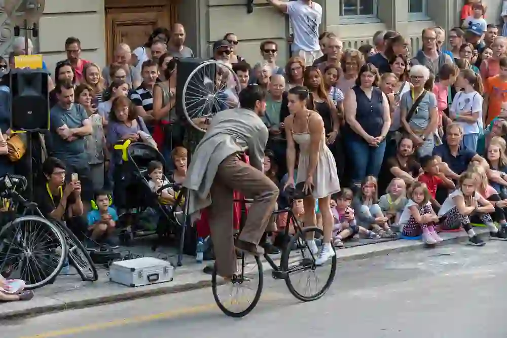 Ein Strassenkünstler auf einem Hochrad fesselt das Publikum beim Buskers Festival in Bern. Die Zuschauer, Jung und Alt, blicken gespannt zu, während ein Helfer die Show unterstützt. Man spürt die Neugier und Begeisterung in den Gesichtern der Menge, die in dichten Reihen um die Szene herum steht. Ein Moment des Staunens und der Freude, der die spielerische und lebendige Atmosphäre des Strassenfestivals einfängt.