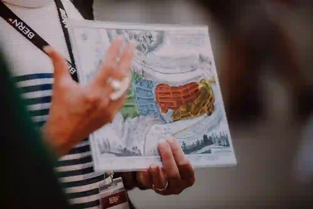 Das Bild zeigt eine Nahaufnahme der Hände einer Person, die eine historische Karte von Bern hält. Die Karte ist bunt und detailliert, markiert verschiedene Stadtteile in unterschiedlichen Farben. Die Person, vermutlich ein Stadtführer, deutet auf einen bestimmten Abschnitt der Karte, möglicherweise um die Aufmerksamkeit der Zuhörer auf einen historisch wichtigen Ort oder ein Ereignis zu lenken. Der Fokus auf die Karte und die gestikulierende Hand vermitteln ein Gefühl von Wissbegierde und Entdeckung.