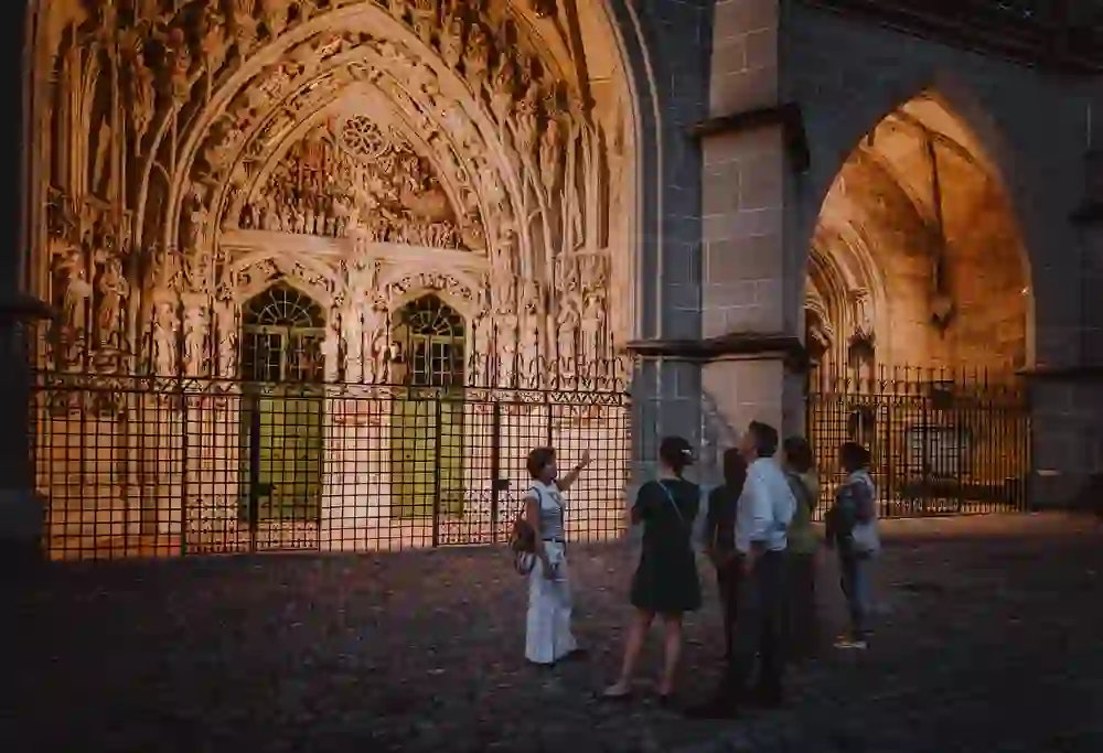 Auf dem Bild ist eine Gruppe von Menschen bei einer abendlichen Stadtführung zu sehen. Sie stehen vordem beeindruckenden Münster Kirchentor, erleuchtet durch warmes Licht, das die filigranen Steinfiguren und Verzierungen hervorhebt. Die Stimmung wirkt ruhig und nachdenklich, als würden die Besucher die historische Schönheit und die Atmosphäre der Umgebung in sich aufnehmen.
