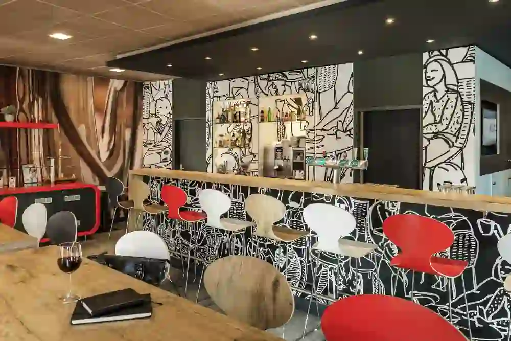 The picture shows the modern restaurant of the Hotel Ibis Expo Bern with a lively atmosphere, characterised by a bar with an eye-catching black and white wall drawing and red accents. Stylish chairs and tables blend in with the modern design. A glass of red wine and a notebook on a wooden table suggest a relaxed environment that invites you to linger.