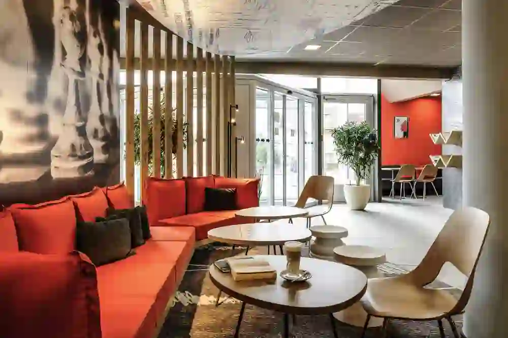 The picture shows a stylish lounge area of the Hotel Ibis Bern Expo with vibrant red sofas and cushions, wooden tables and modern chairs. A ceiling with sketches hangs above the area and a chess piece adorns one wall. The atmosphere is modern, comfortable and inviting.