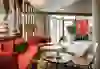 L'immagine mostra un'elegante area lounge dell'Hotel Ibis Bern Expo con divani e cuscini di colore rosso vivo, tavoli in legno e sedie moderne. Un soffitto con schizzi è appeso sopra l'area e un pezzo di scacchi adorna una parete. L'atmosfera è moderna, confortevole e invitante.