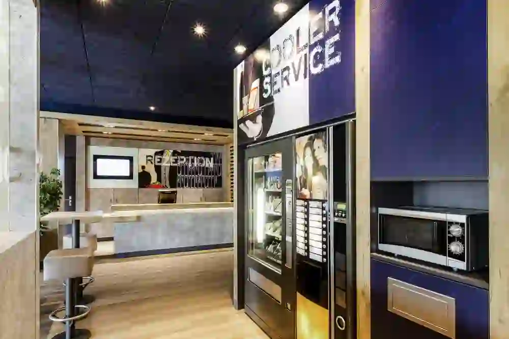 The photo shows the reception area of the Ibis Budget Bern Expo hotel with a modern, sober design. A snack and drinks vending machine stands next to a microwave for guests to serve themselves. The area is well-lit, clean and functional, with a relaxed and welcoming atmosphere.