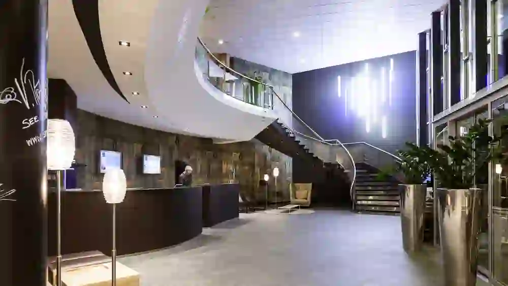 L'immagine mostra la hall del Novotel Bern Expo dal design moderno ed elegante. La scala curva e l'illuminazione dinamica creano un senso di movimento e di comfort contemporaneo che invita i visitatori in un mondo di moderna ospitalità.