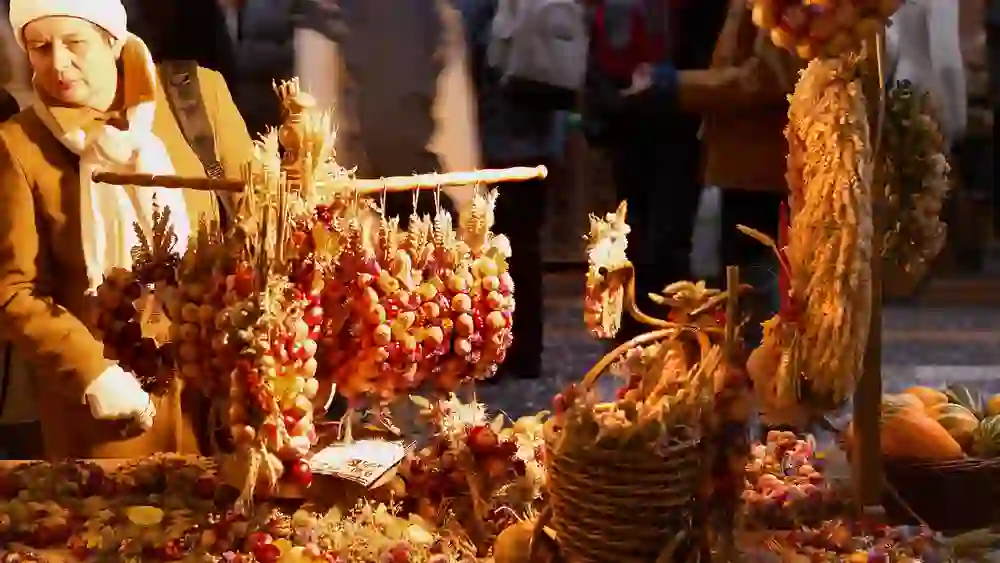 Das Bild zeigt einen belebten Marktstand auf Zibelemärit (Zwiebelmarkt). Im Vordergrund sind verschiedene dekorative Kränze und Gestecke aus Zwiebeln und getrockneten Blumen zu sehen. Eine Frau mit weißer Mütze und Schal betrachtet die ausgestellten Waren aufmerksam. Der Hintergrund ist verschwommen, aber weitere Marktbesucher und Stände sind erkennbar, was auf eine geschäftige Atmosphäre hinweist. Das warme Licht verstärkt die gemütliche Stimmung des Marktes.
