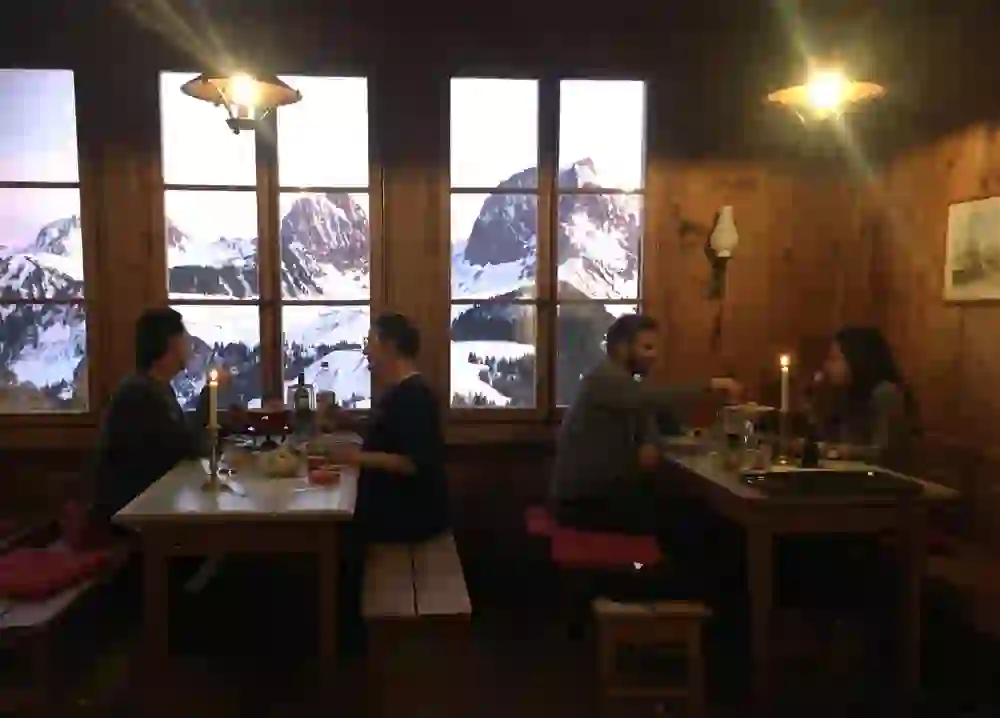 Das Bild zeigt zwei Paare, die in einer gemütlichen Holzhütte bei Kerzenlicht ein Fondue genießen. Durch die Fenster sieht man eine atemberaubende winterliche Berglandschaft. Die warme, intime Atmosphäre vermittelt ein Gefühl von Geborgenheit und Zusammengehörigkeit.