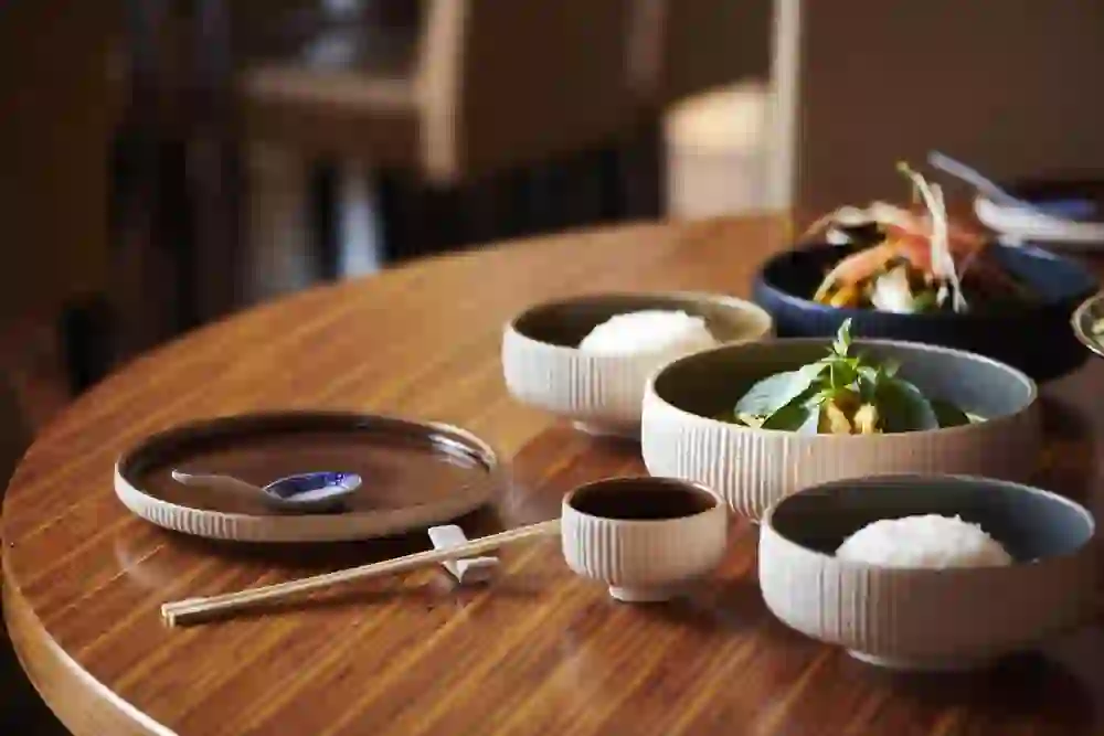 The picture shows a dining table at the Mishio Vatterland restaurant in Bern. There are various dishes that appear to have an Asian flavour - including two bowls of white rice, a bowl of green vegetables and basil leaves, and a bowl of a vegetable and meat dish, presumably a curry or similar stew. There are also chopsticks and small bowls, presumably for soya sauce, on the table.