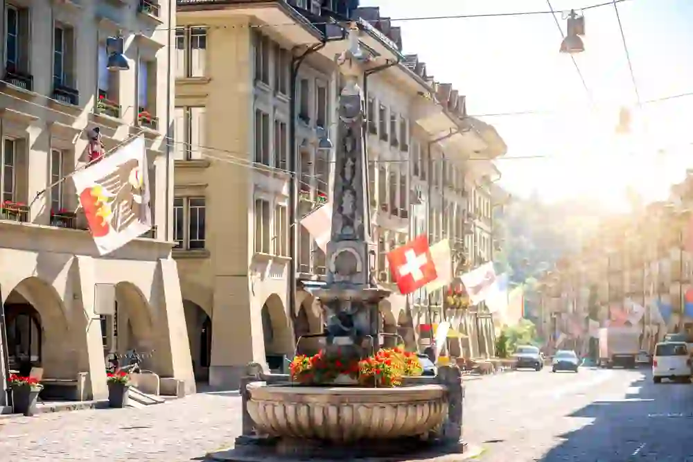 Die Gerechtigkeitsgasse in Bern, geschmückt mit Schweizer Fahnen und Blumen am Brunnen, badet im goldenen Licht eines klaren Morgens, ein Bild der Ruhe und des Stadtlebens in Harmonie.