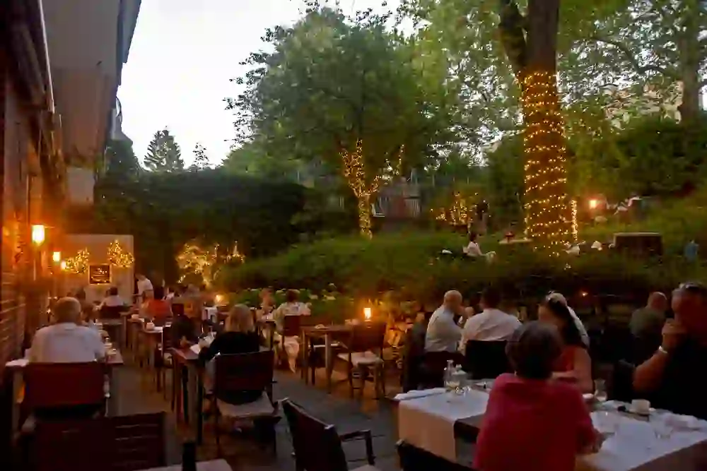 The photo shows a lively scene in the outdoor area of the Essort restaurant in Bern at dusk. The tables are elegantly laid and guests are enjoying their meals. Fairy lights on the trees create a magical atmosphere. The natural surroundings and soft lighting create a relaxing and romantic ambience