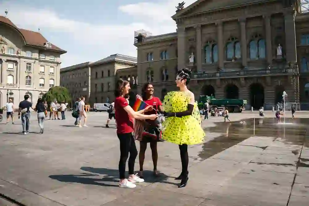 A radiant drag queen in a striking yellow dress chats happily with two people in front of the Federal Palace, children play in the background.