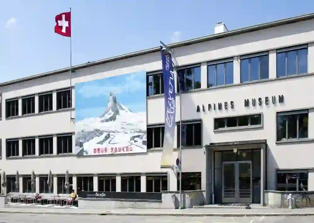 The photo shows the Alpine Museum in sunny weather, which inspires cheerfulness and interest in the mountain world. The Swiss flag flies proudly next to a vivid image of the Matterhorn.
